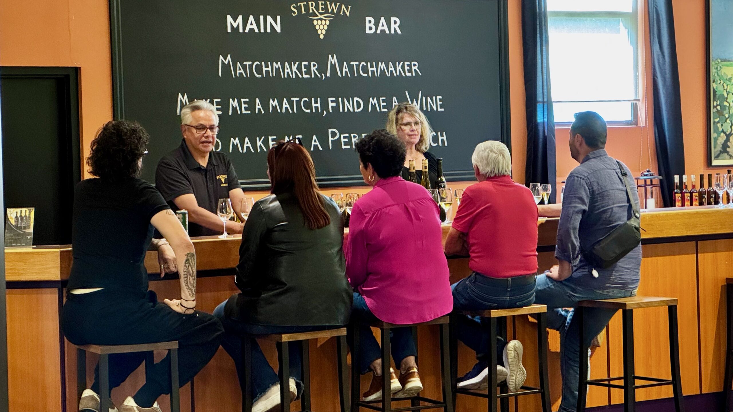 Group of people enjoying wine tasting at Strewn Winery bar with a chalkboard menu in the background.