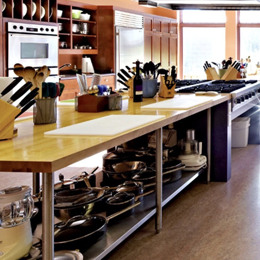 Cooking school kitchen with wooden cabinets, stainless steel appliances, and a large island with cooking utensils and cookware.