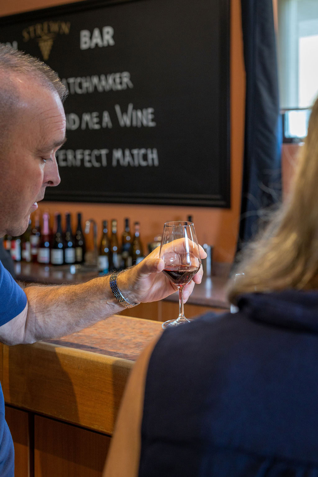 Man tasting red wine at Strewn Winery with bar and wine bottles in background.